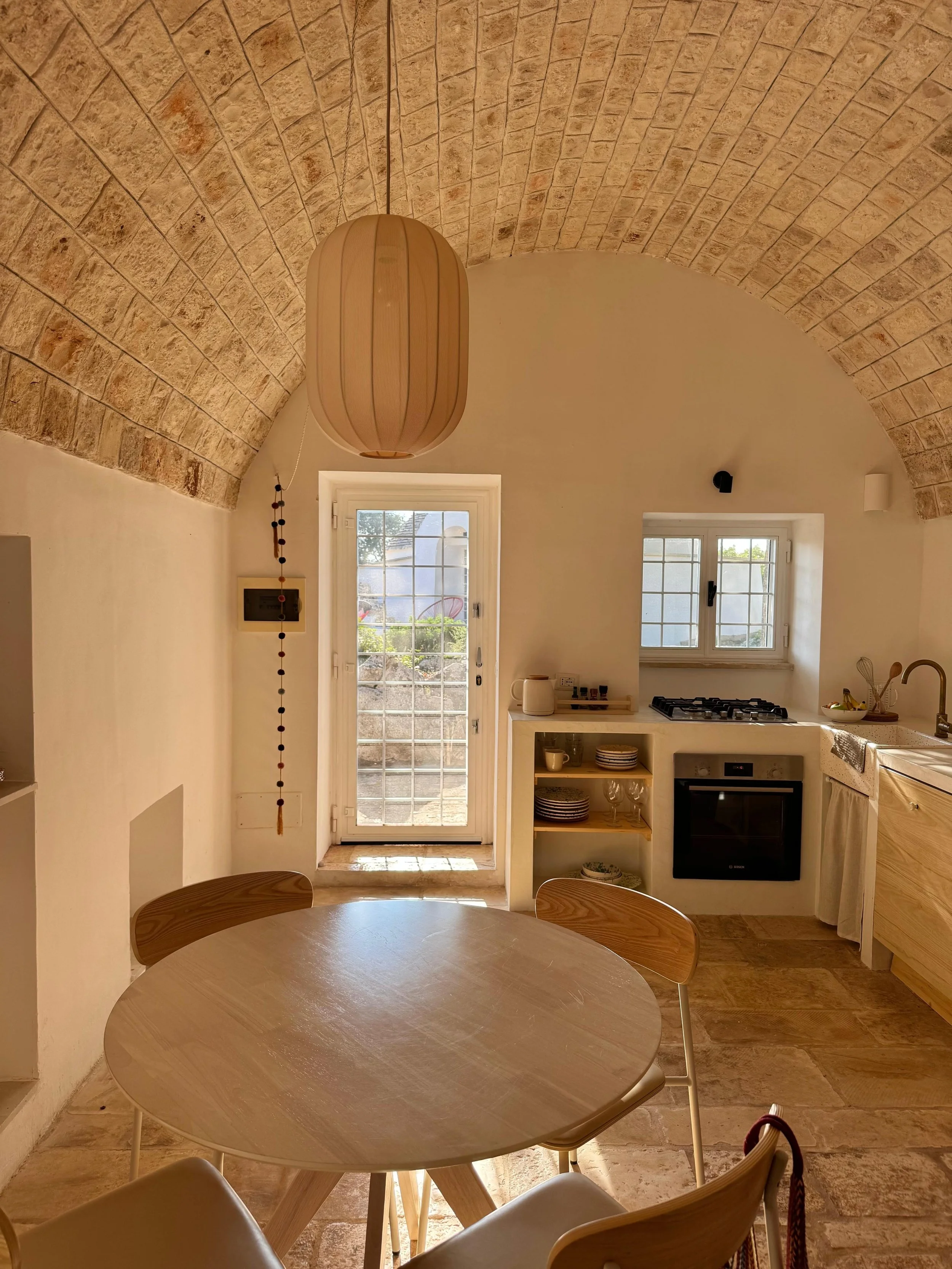 Binnenkijkje van een keuken met een houten eettafel en stoelen, een deur met ramen die uitkijken op een tuin, een raam boven het aanrecht, een hanglamp, en keukenspullen op planken.