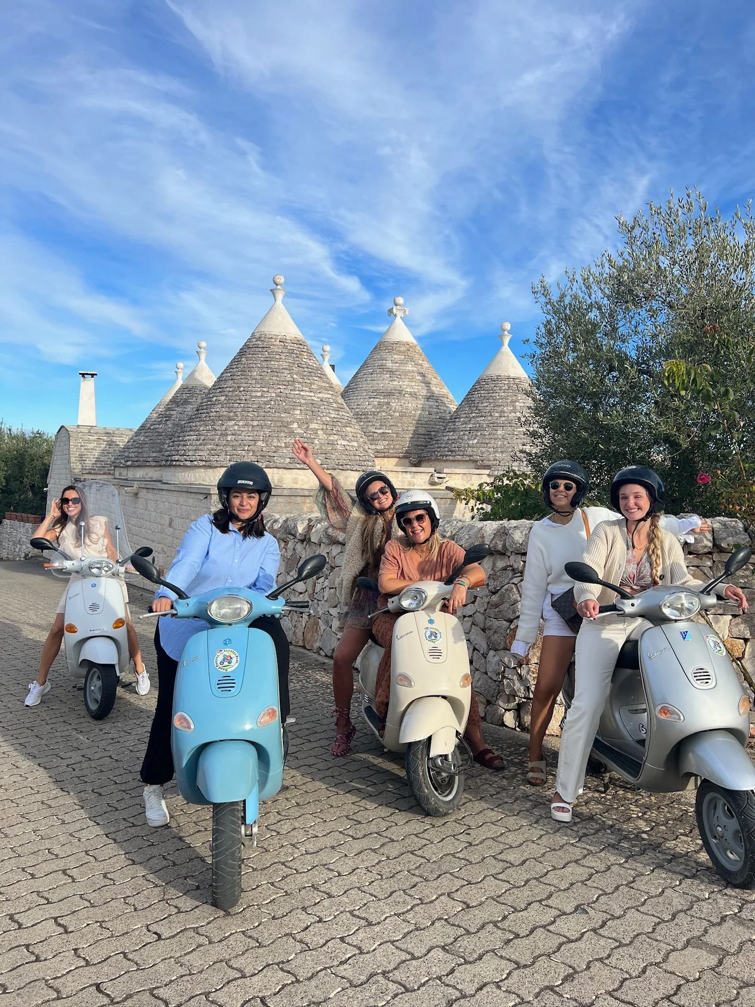 Vijf vrouwen op scooters poseert voor een stenen muur onder een blauwe hemel met enkele wolken, met traditionele trulli-gebouwen op de achtergrond.