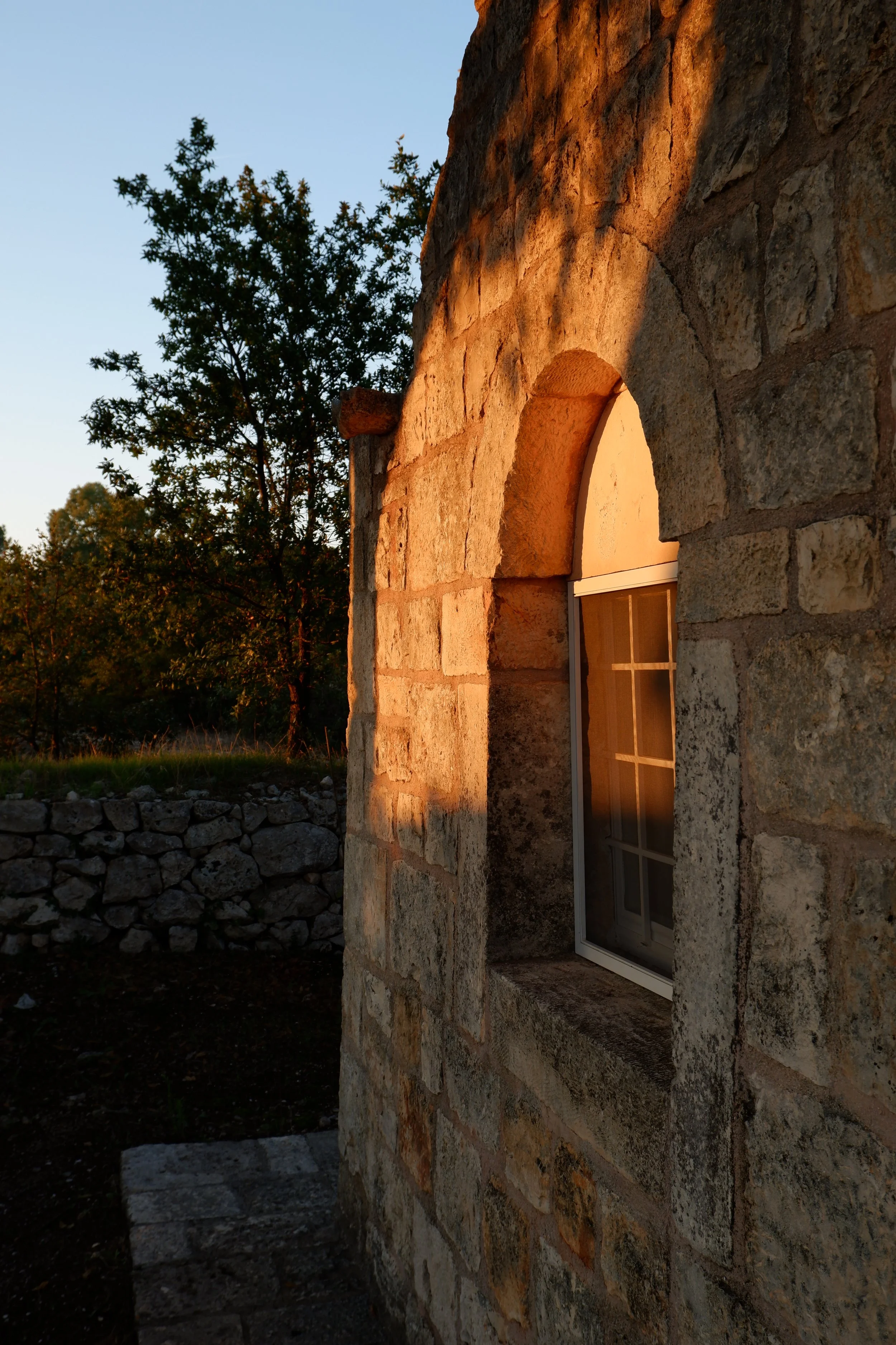 Een stenen muur met een klein raampje dat door de zon verlicht wordt, met een achtergrond van bomen onder een heldere hemel.