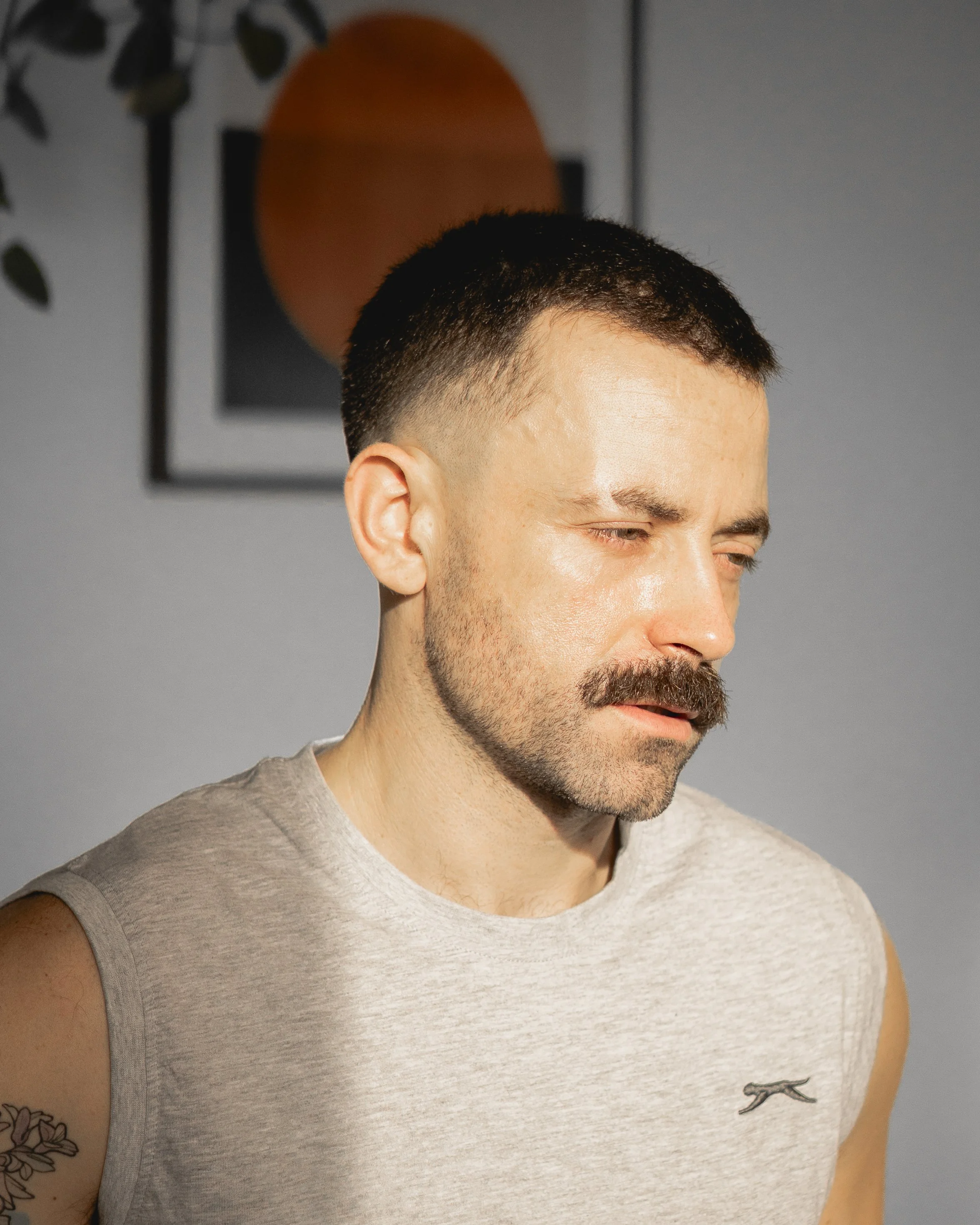 A gay man with a mustache, short dark hair, wearing a sleeveless gray shirt, looking down, in front of a wall with modern artwork, ready to give a gay massage.