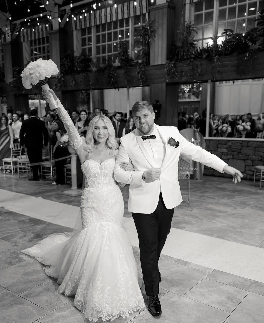A newlywed couple celebrating their wedding indoors, with the bride holding a bouquet raised in the air and wearing a lace wedding gown, and the groom in a white tuxedo with a bow tie, both smiling and waving to the camera, in front of seated guests 