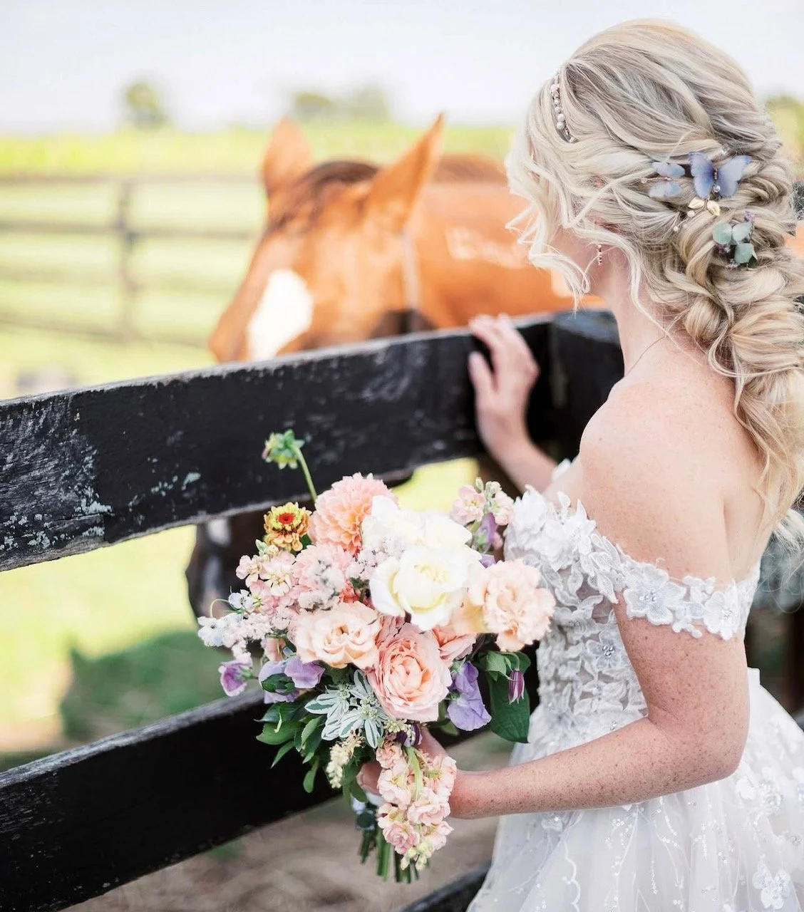 A bride with blonde hair decorated with blue and silver hair accessories, wearing a white lace wedding dress, holding a bouquet of pink, purple, and white flowers, stands by a wooden fence and looks at a brown horse in a field.