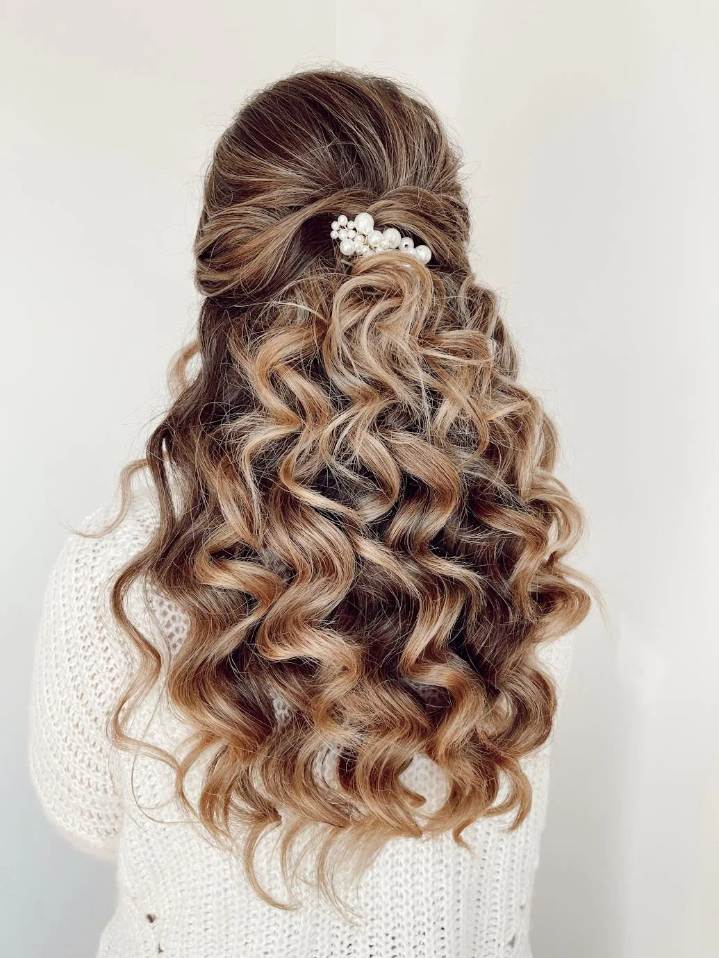 Back view of a woman with long, curly, blonde hair adorned with a white pearl hair clip.