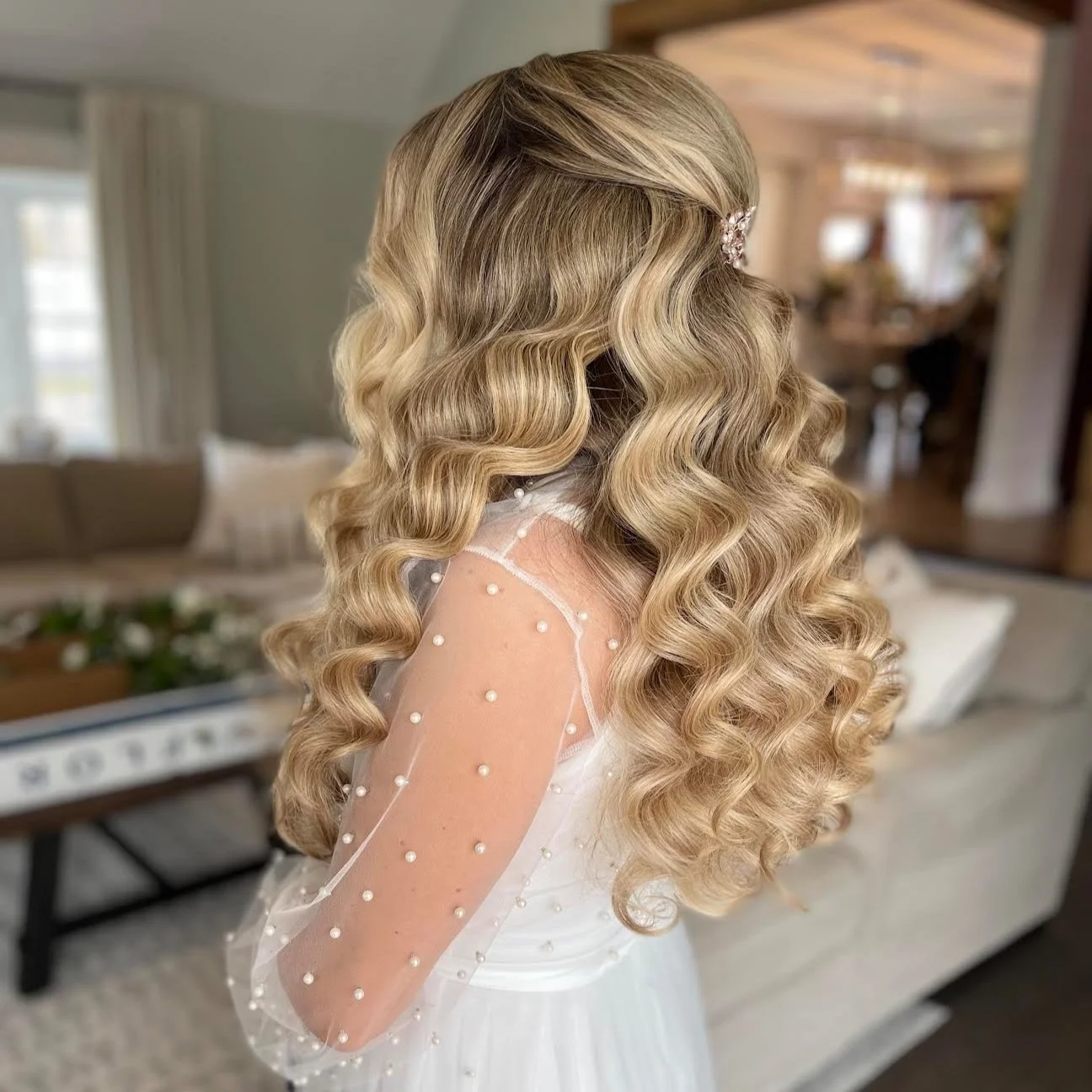 A woman with long, blonde, wavy hair styled in loose curls, wearing a white dress with sheer, beaded sleeves, sitting in a cozy, well-lit living room.