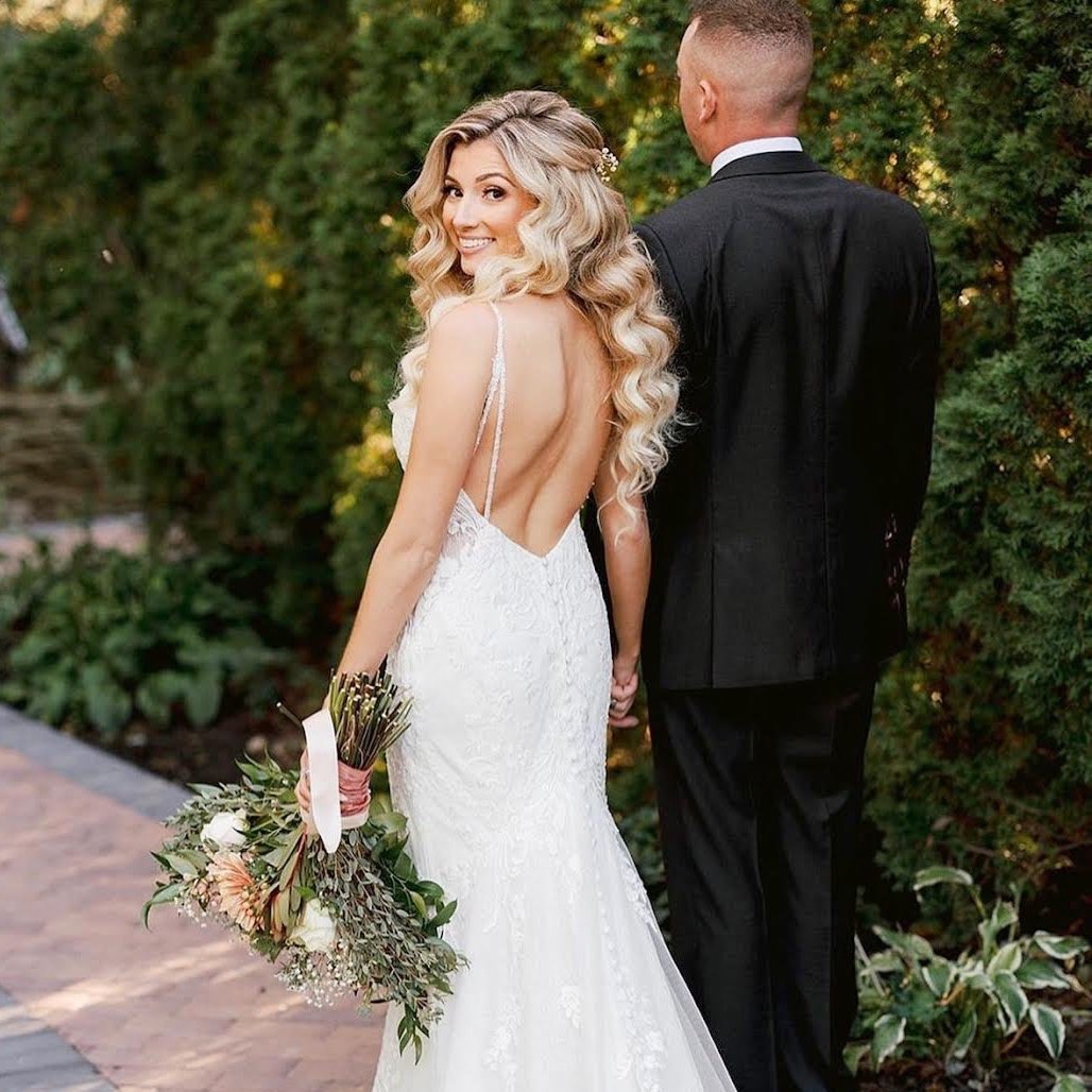 A bride in a white lace wedding gown with spaghetti straps and backless design, holding a bouquet of flowers, smiling over her shoulder, standing outdoors with a groom in a black suit and white shirt, against a background of green shrubs.