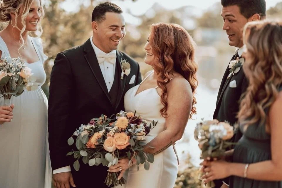 A bride and groom smiling at each other during a wedding ceremony, surrounded by bridesmaids and groomsmen holding bouquets, outdoors near water.