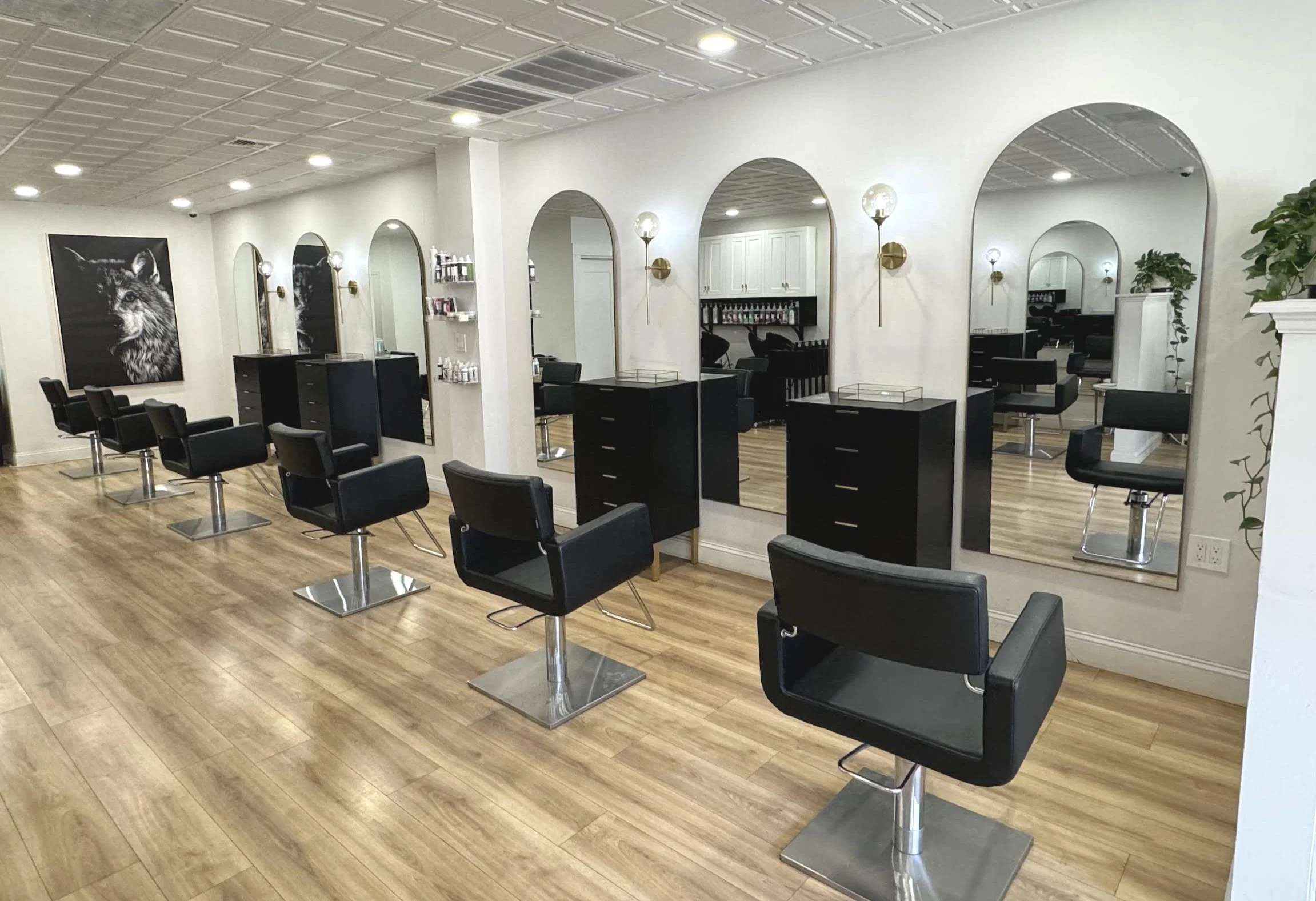 Empty hair salon with black chairs, mirrors, wooden flooring, and wall-mounted lights.