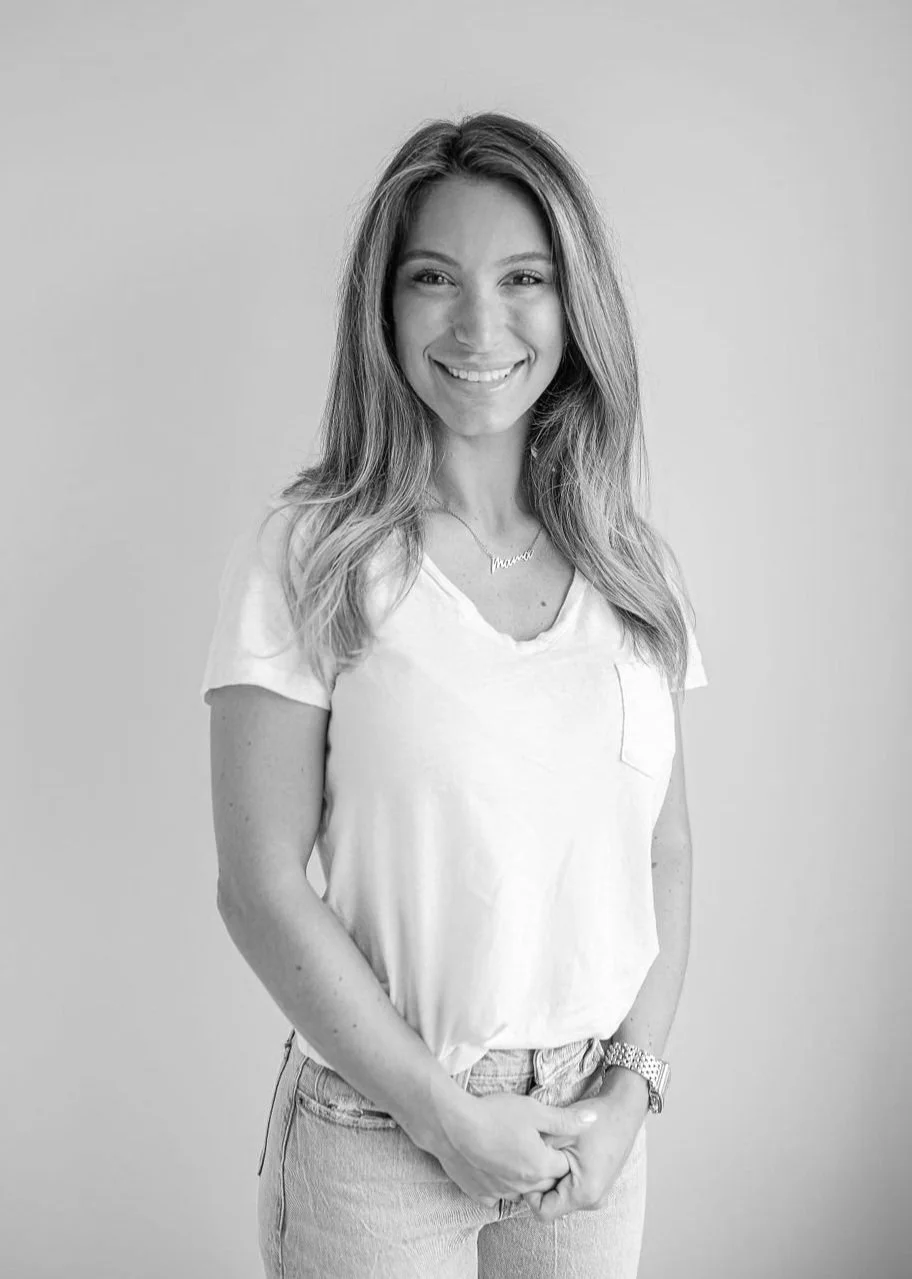 A smiling woman with long wavy hair wearing a light-colored t-shirt and jeans, standing against a plain background.