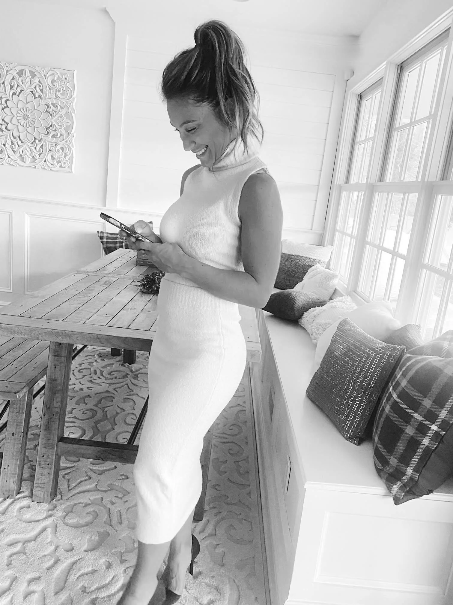 A woman with a high ponytail wearing a sleeveless dress and high heels is looking at her phone, smiling, in a well-lit room with large windows, a wooden dining table, and some pillows on a window seat.