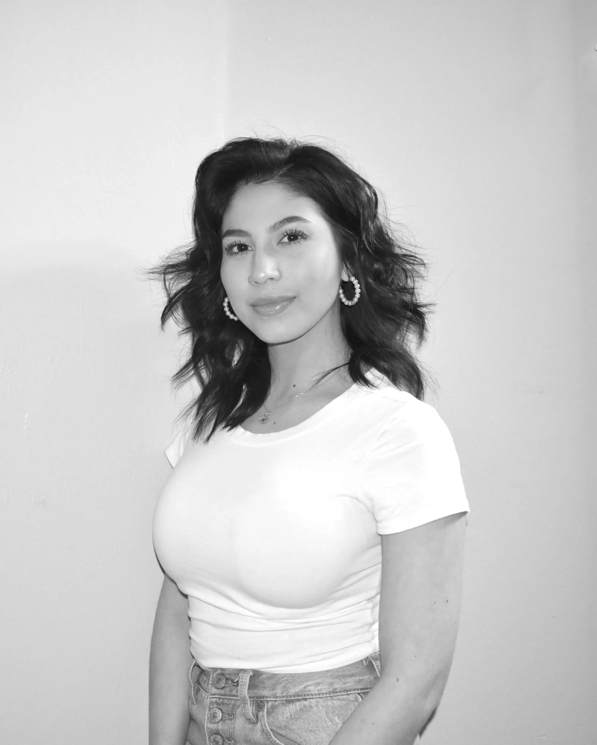 Black and white photo of a young woman with wavy dark hair, wearing a white t-shirt, hoop earrings, and a necklace, standing against a plain wall.