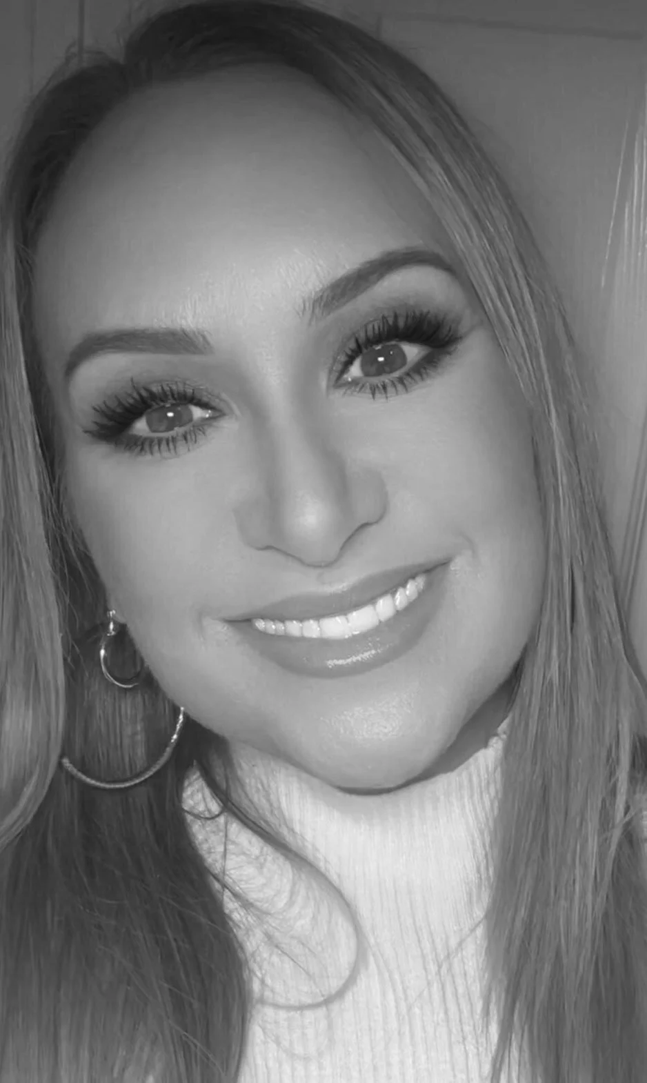 A close-up black and white portrait of a smiling woman with long hair, wearing hoop earrings and a light-colored turtleneck sweater.
