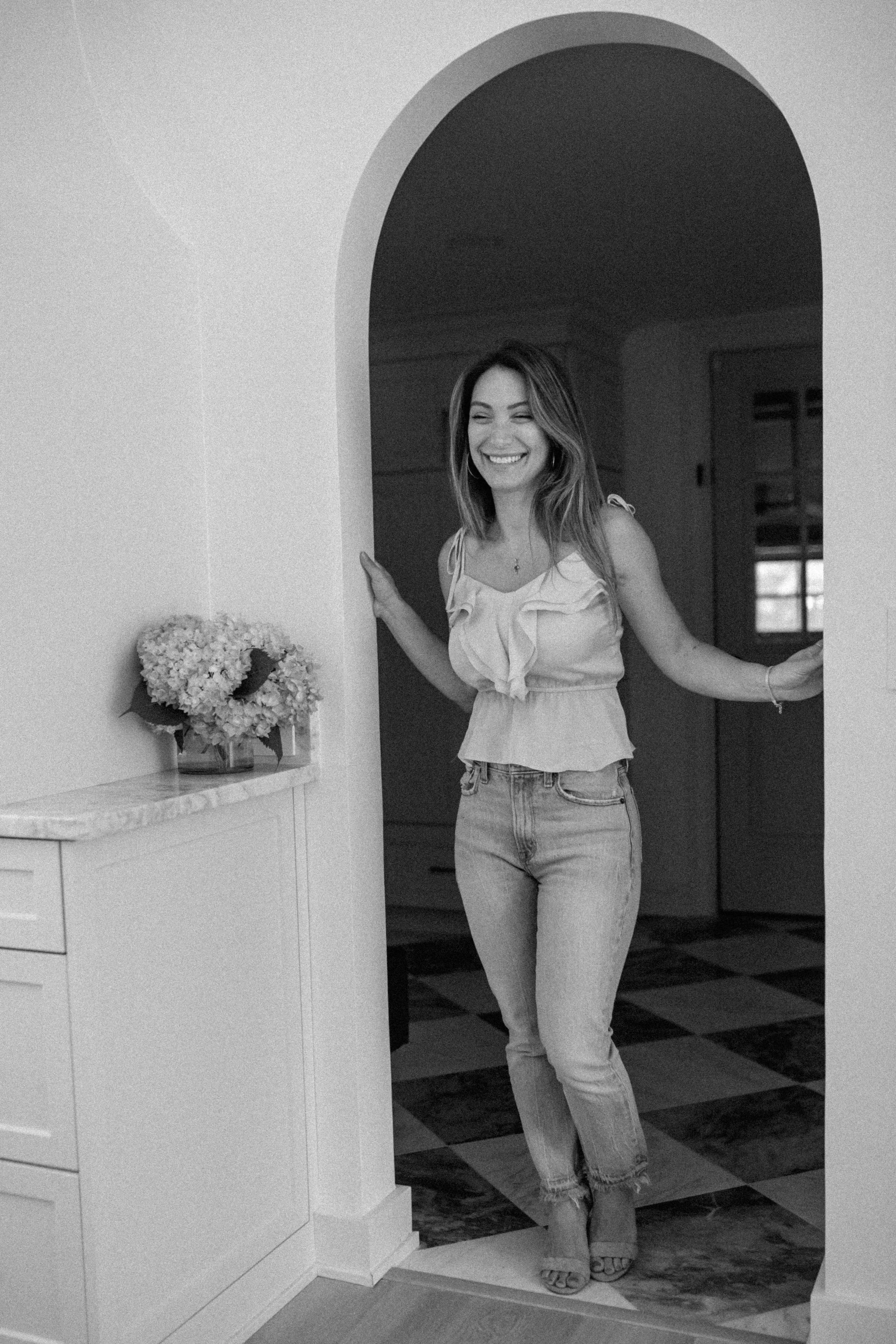 A woman standing in a doorway, smiling and looking down. She is wearing a sleeveless ruffled top and jeans. There is a vase of flowers on a counter to her left.