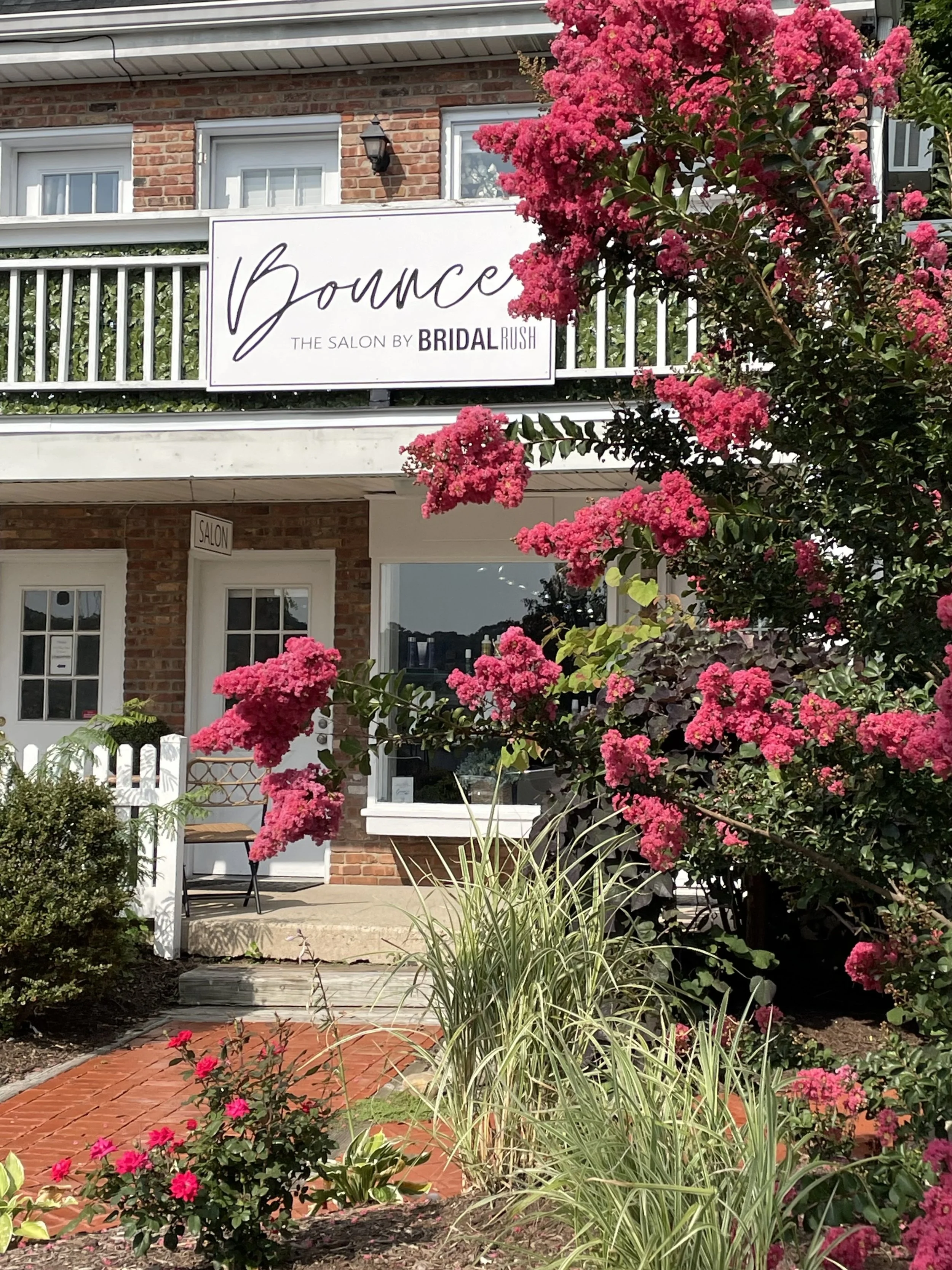 The exterior of a salon named Bounce, with brick walls, a white sign, and a porch. Bright pink flowering bushes, green plants, and a red brick pathway leading to the entrance.