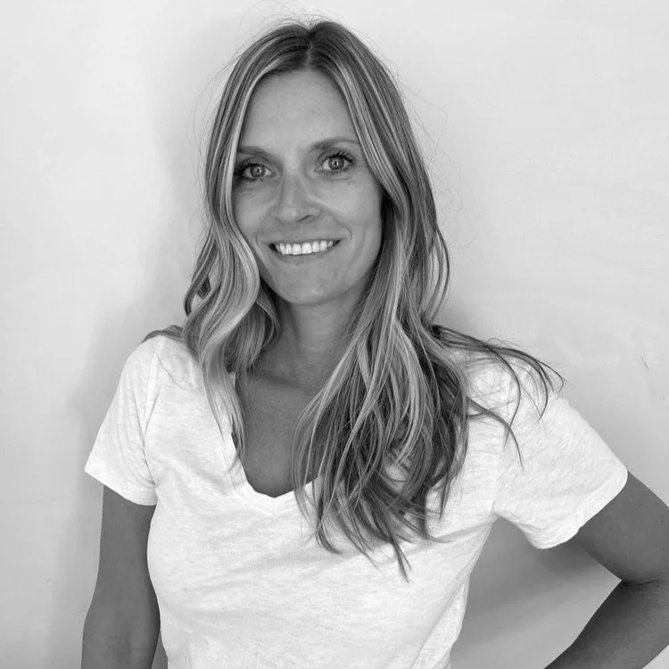 A woman with long wavy hair smiling at the camera, wearing a white t-shirt against a plain white background.