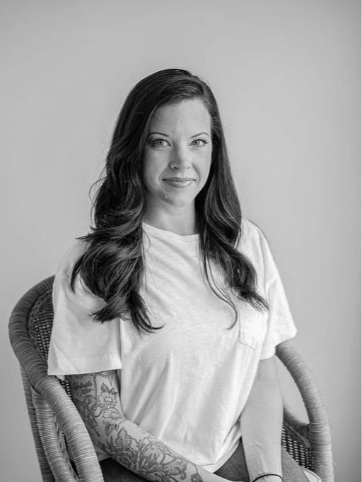 Black and white photo of a woman with long wavy hair, wearing a casual t-shirt, sitting on a wicker chair with a tattooed arm, smiling softly.