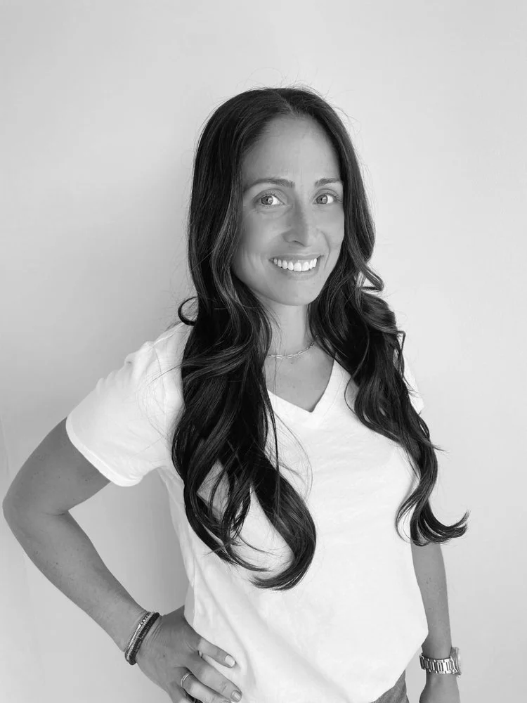 Black and white photo of a woman with long wavy dark hair, smiling, wearing a white t-shirt, a wristwatch, bracelet, and a necklace, standing against a plain wall.