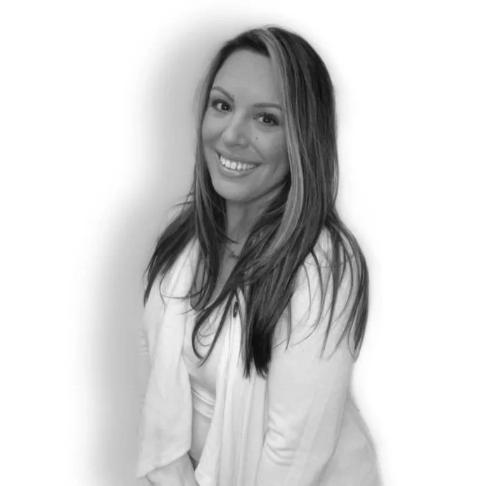 A woman with long hair smiling and looking at the camera, wearing a light-colored top or jacket, in front of a plain white background.