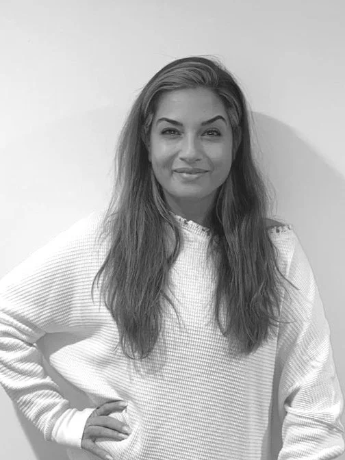 A woman with long, wavy hair, wearing a white sweater, standing against a plain wall, smiling, with one hand on her hip.
