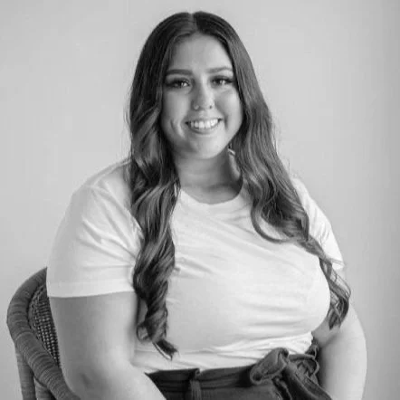 Black and white photo of a young woman with long wavy hair, smiling, wearing a casual white t-shirt, sitting on a chair.