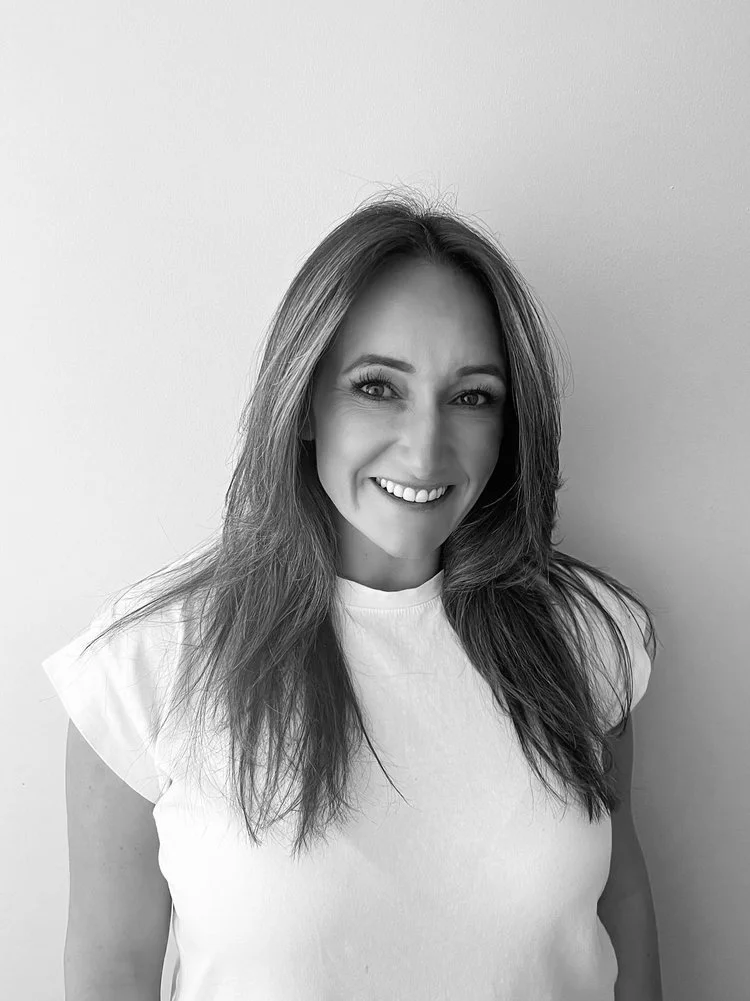 A woman with long, wavy hair smiling and looking at the camera, wearing a white t-shirt, standing against a plain light-colored wall.
