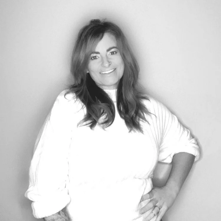 Smiling woman with wavy hair wearing a light-colored, long-sleeved top, standing against a plain background.