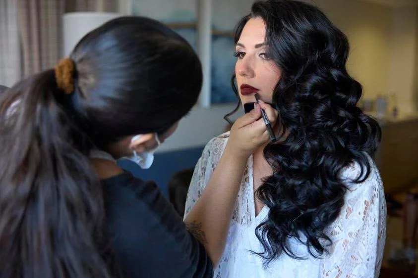 Makeup artist applying lipstick to a woman with long black curly hair and lace top in a room.