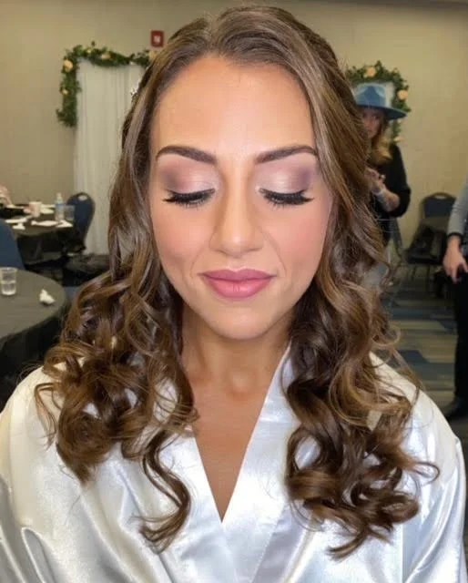 A woman with long curly brown hair and makeup, wearing a white satin robe, at a social event or celebration in a decorated room.