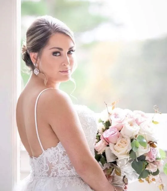 Bride in a wedding dress holding a bouquet of pink and white roses, standing by a window.