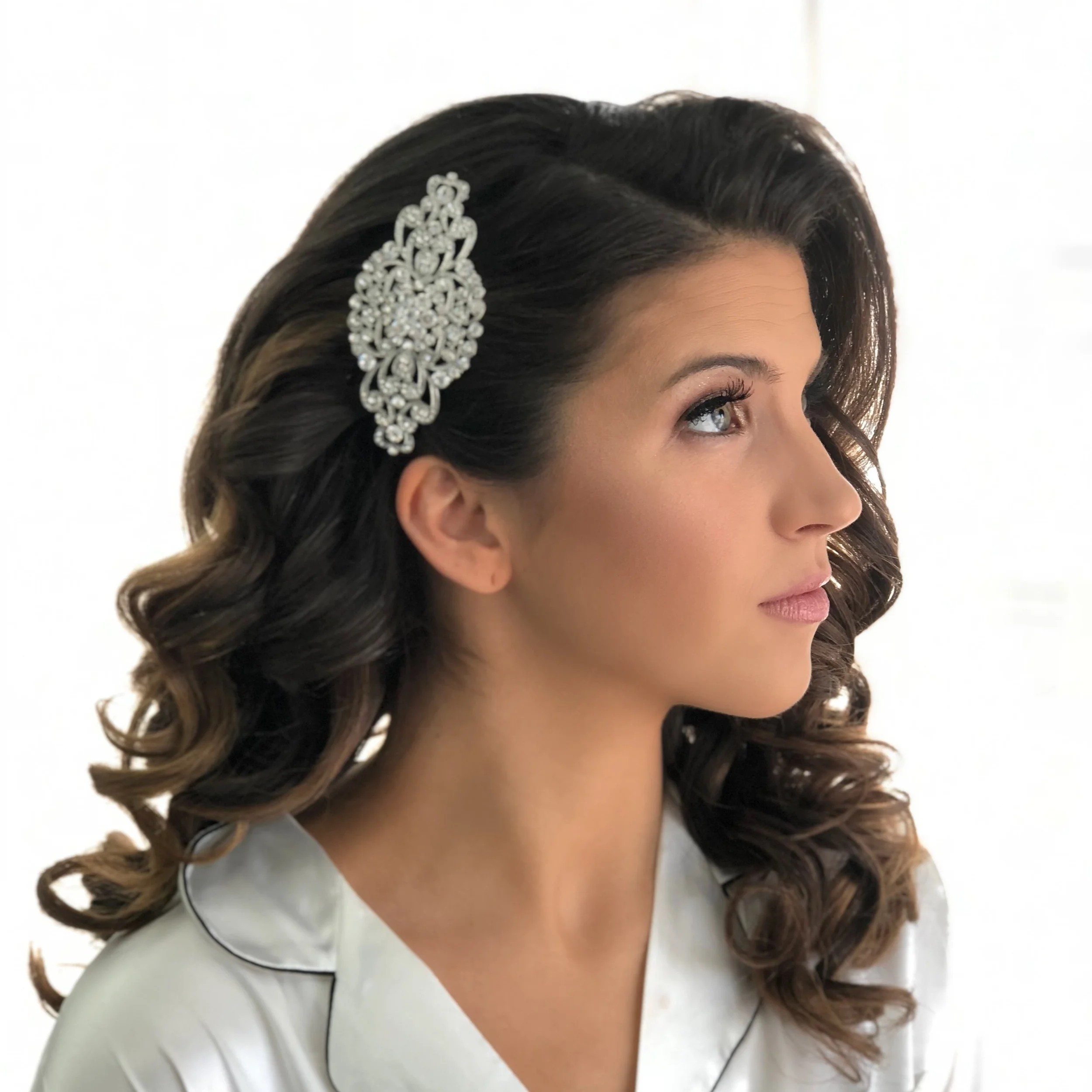 Side profile of a woman with medium-length wavy brown hair adorned with a large, ornate silver decorative hairpiece, wearing a white satin robe.