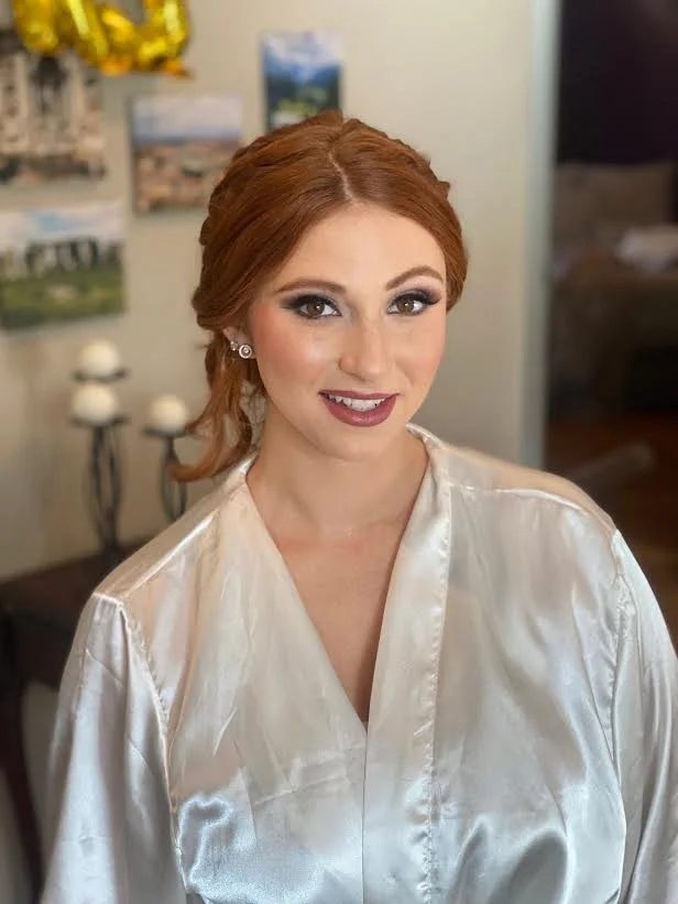 A woman with red hair styled in an elegant updo, wearing makeup with dark eyeshadow, eyeliner, and lipstick, dressed in a white satin robe, standing indoors with blurred background of framed pictures and candles.