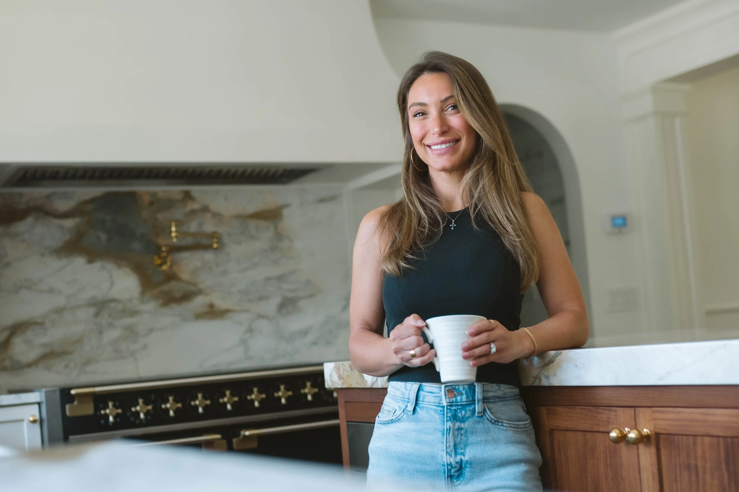 A woman standing in a kitchen, holding a white mug, smiling, wearing a black sleeveless top and light blue jeans.