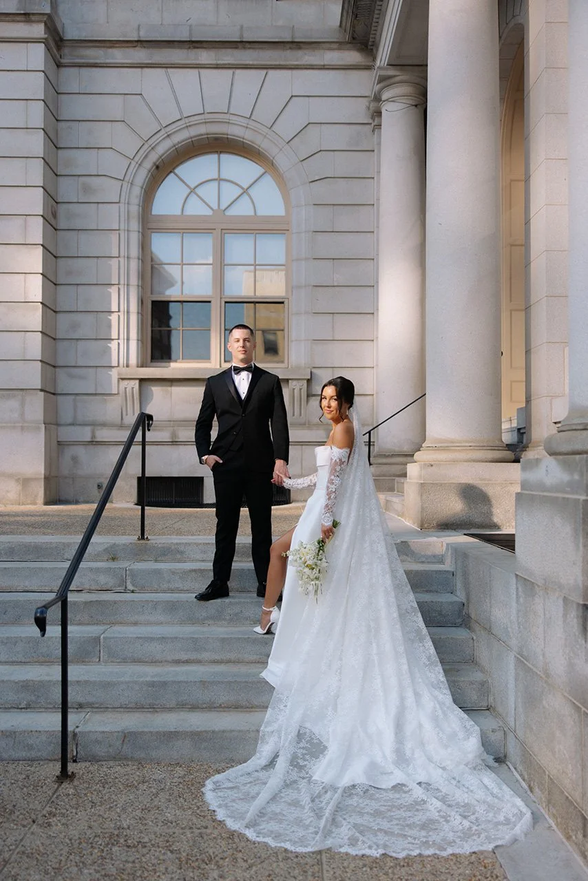 A bride and groom holding hands on steps in front of a neoclassical building with large columns and arched window, during their wedding.