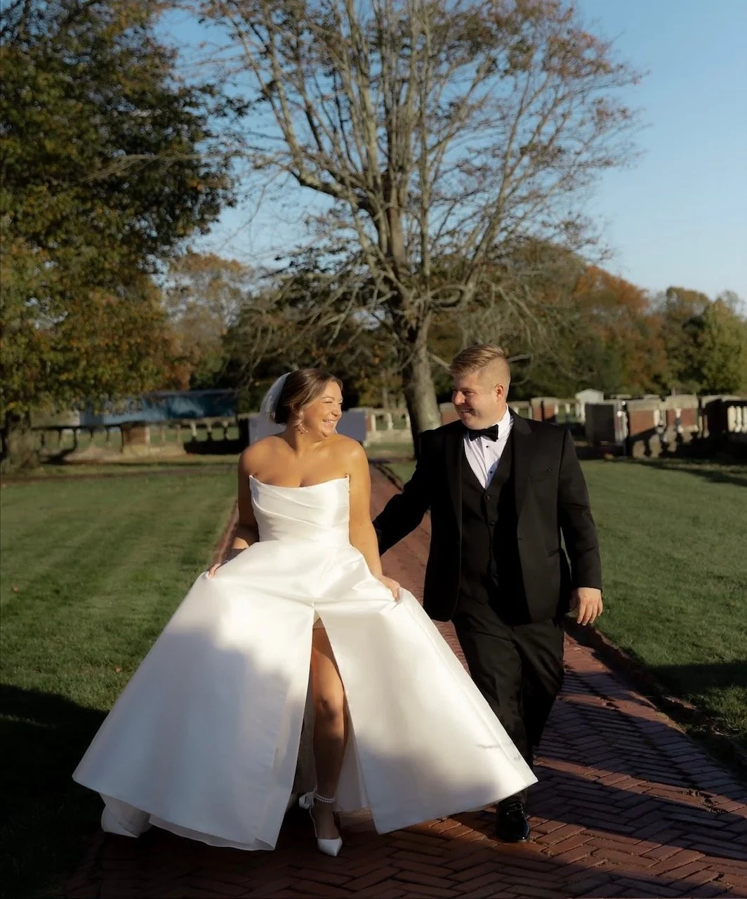 A bride in a white wedding gown and veil and a groom in a black tuxedo walking on a brick path, smiling and holding hands, outdoors with trees and a clear sky.