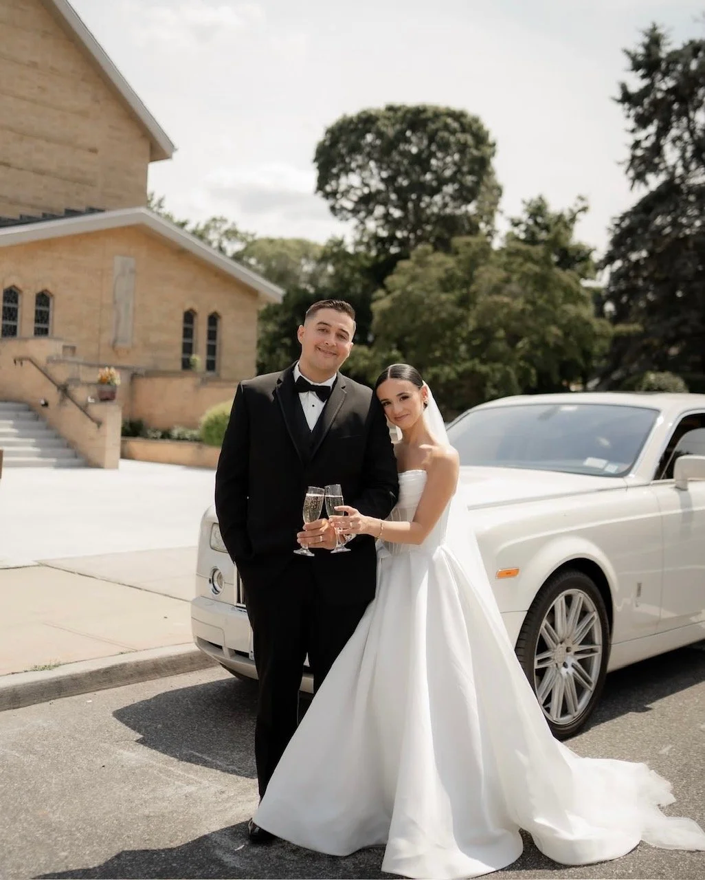 A newlywed couple celebrating outside a church with a white limousine, the groom in a black tuxedo and the bride in a white wedding gown holding champagne glasses.