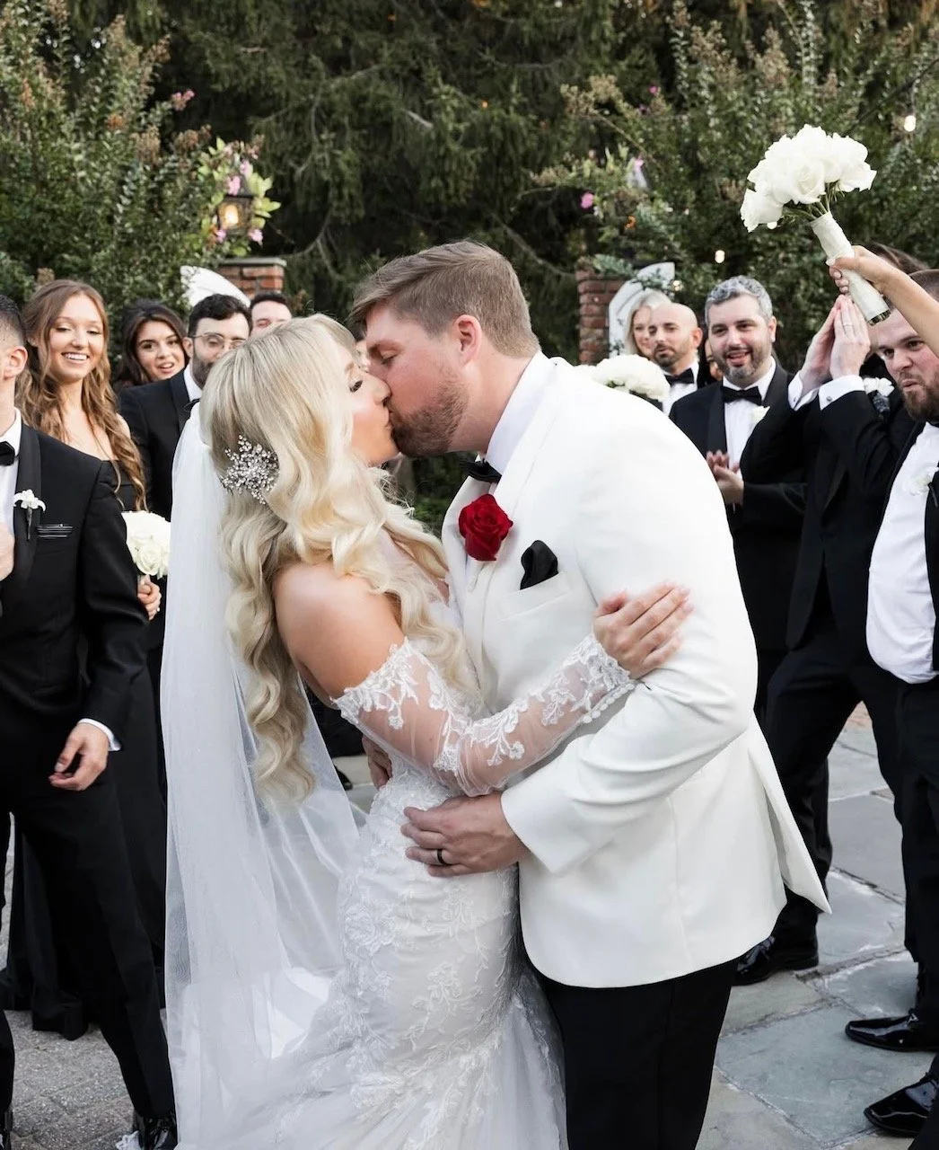 Bride and groom kissing during wedding ceremony surrounded by wedding party and guests outdoors.