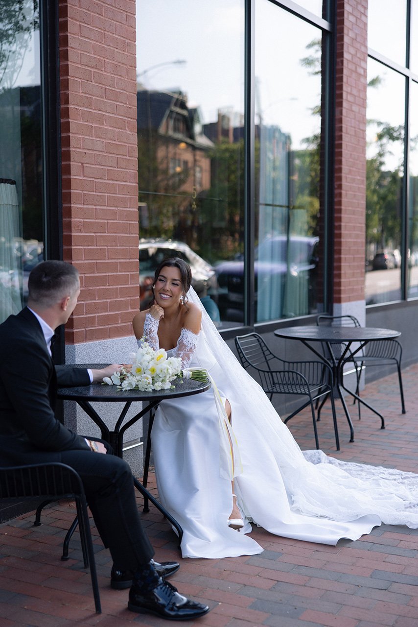 A bride in a white wedding dress sitting at an outdoor cafe table, smiling and looking at a man in a suit, with a bouquet of white flowers on the table.