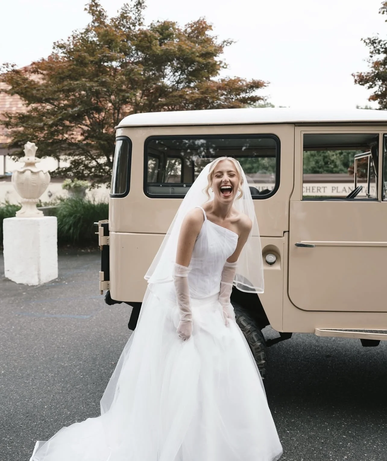 A smiling bride in a white wedding dress and veil, standing outdoors beside a vintage beige vehicle, with trees and a sign in the background.