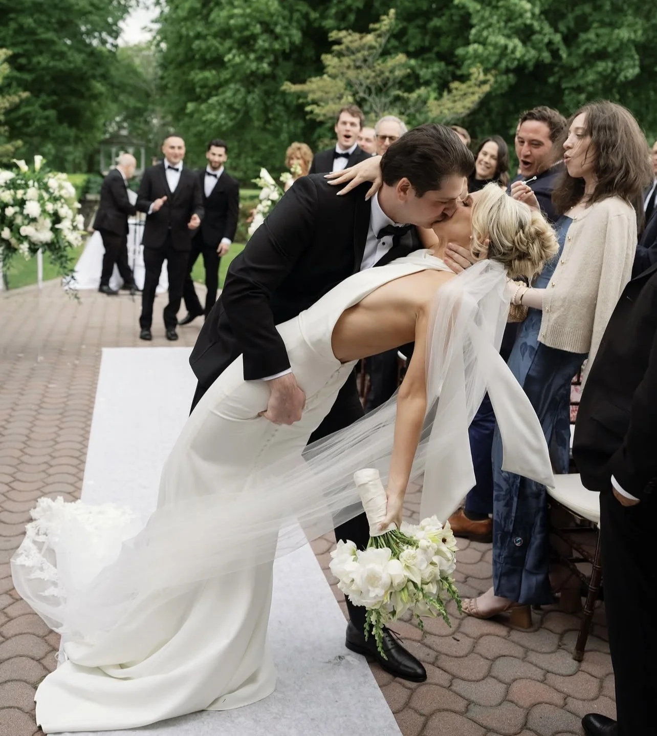 A groom in a black tuxedo kisses a bride in a white wedding dress as she holds a bouquet of white flowers, while wedding guests look on, outdoors with green trees in background.