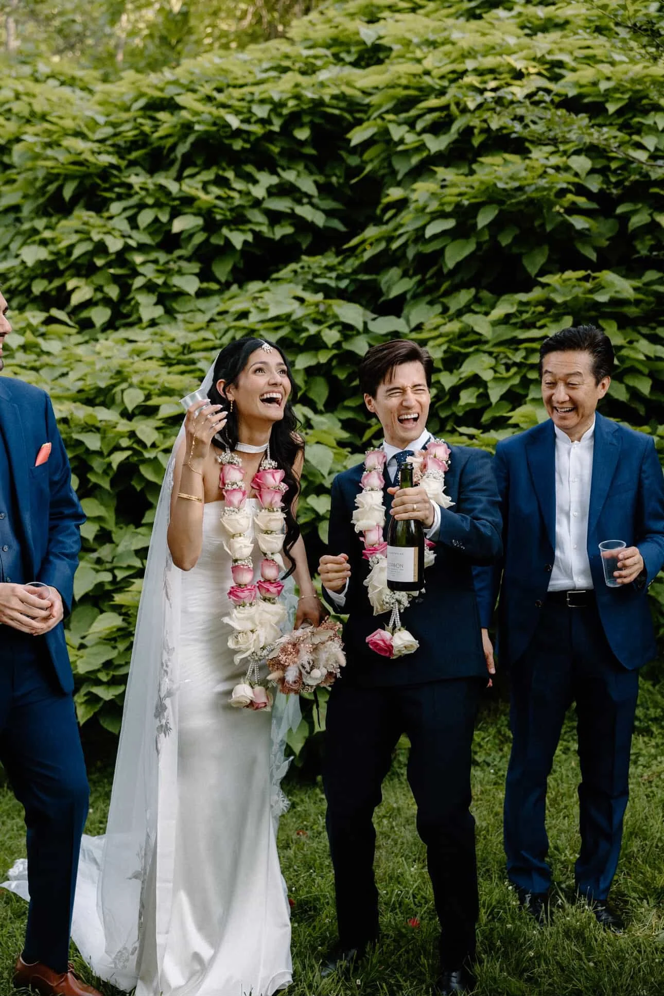 A joyful wedding celebration outdoors with a bride and groom in the center, both wearing leis, surrounded by friends. The bride has long dark hair, a wedding dress, and is holding a bouquet, while the groom is in a dark suit holding a champagne bottl