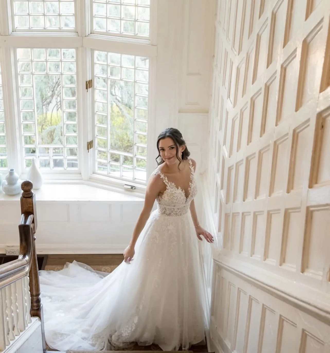Bride in a wedding dress standing on a staircase near a large window with a garden view.