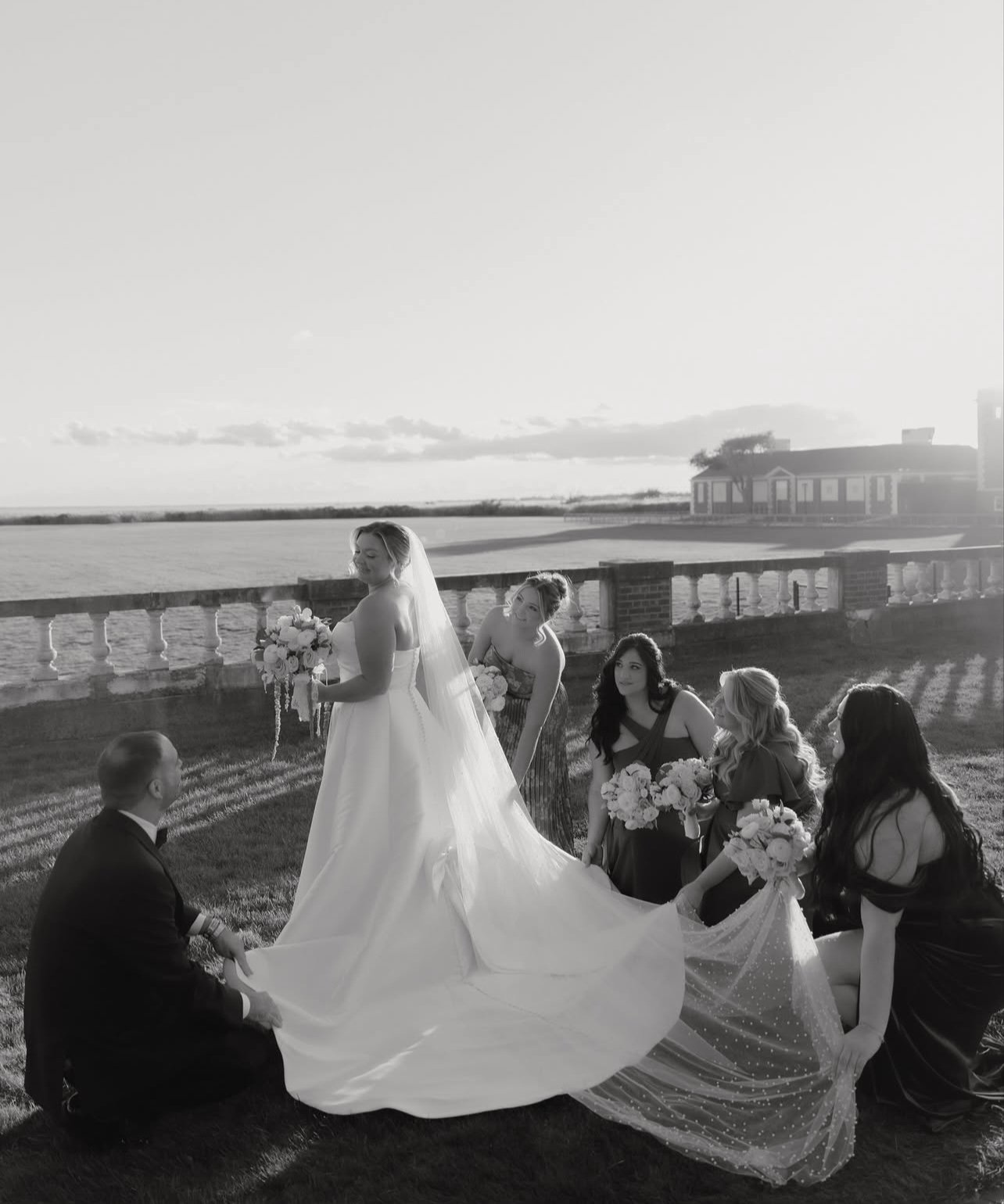 Black and white photograph of a wedding scene outdoors by the water with six people, including a bride in a wedding dress with a veil, a groom kneeling, and four women holding bouquets, during sunset.