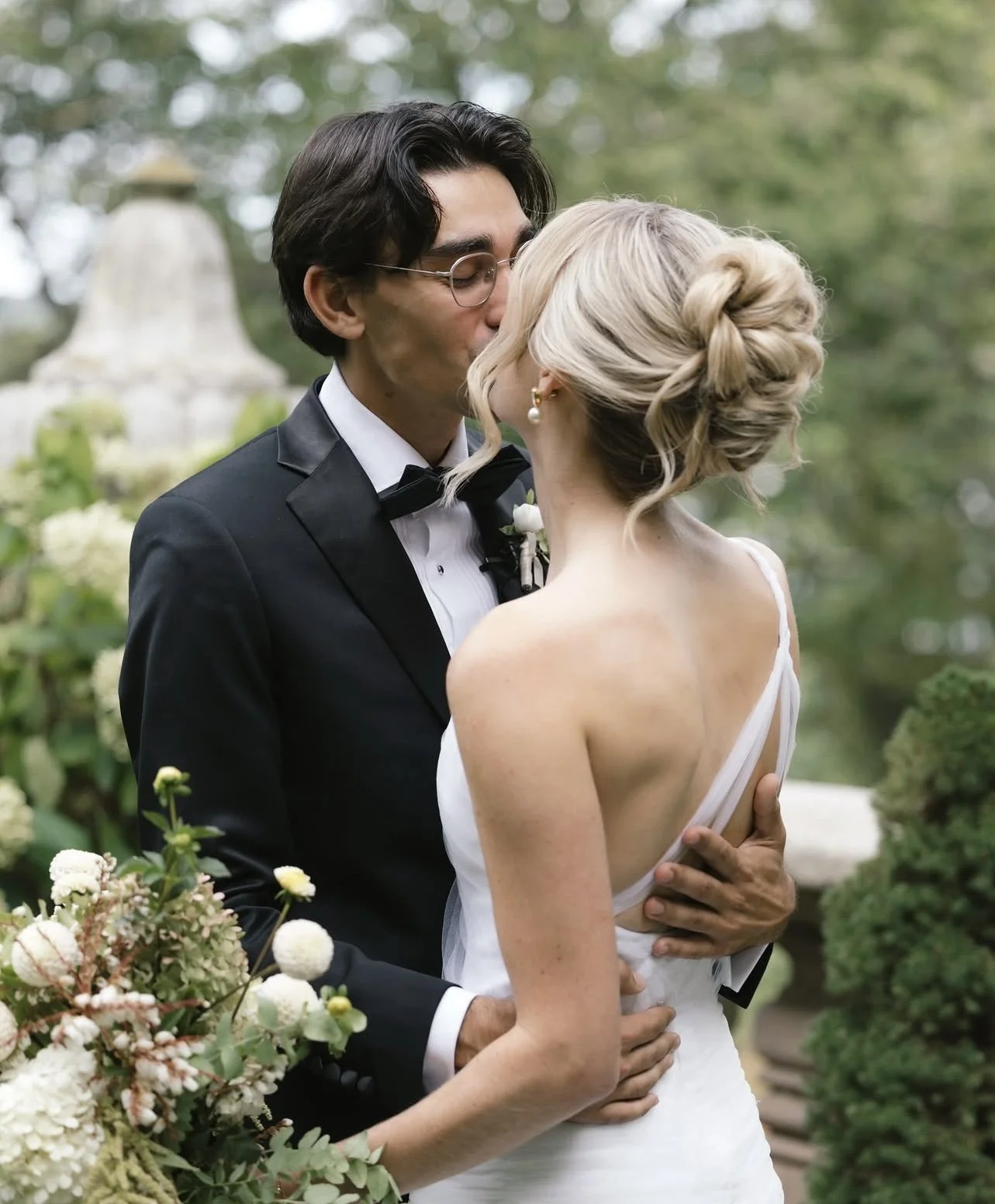 A newlywed couple kissing outdoors, with the bride in a white gown and the groom in a black tuxedo, surrounded by greenery and flowers.