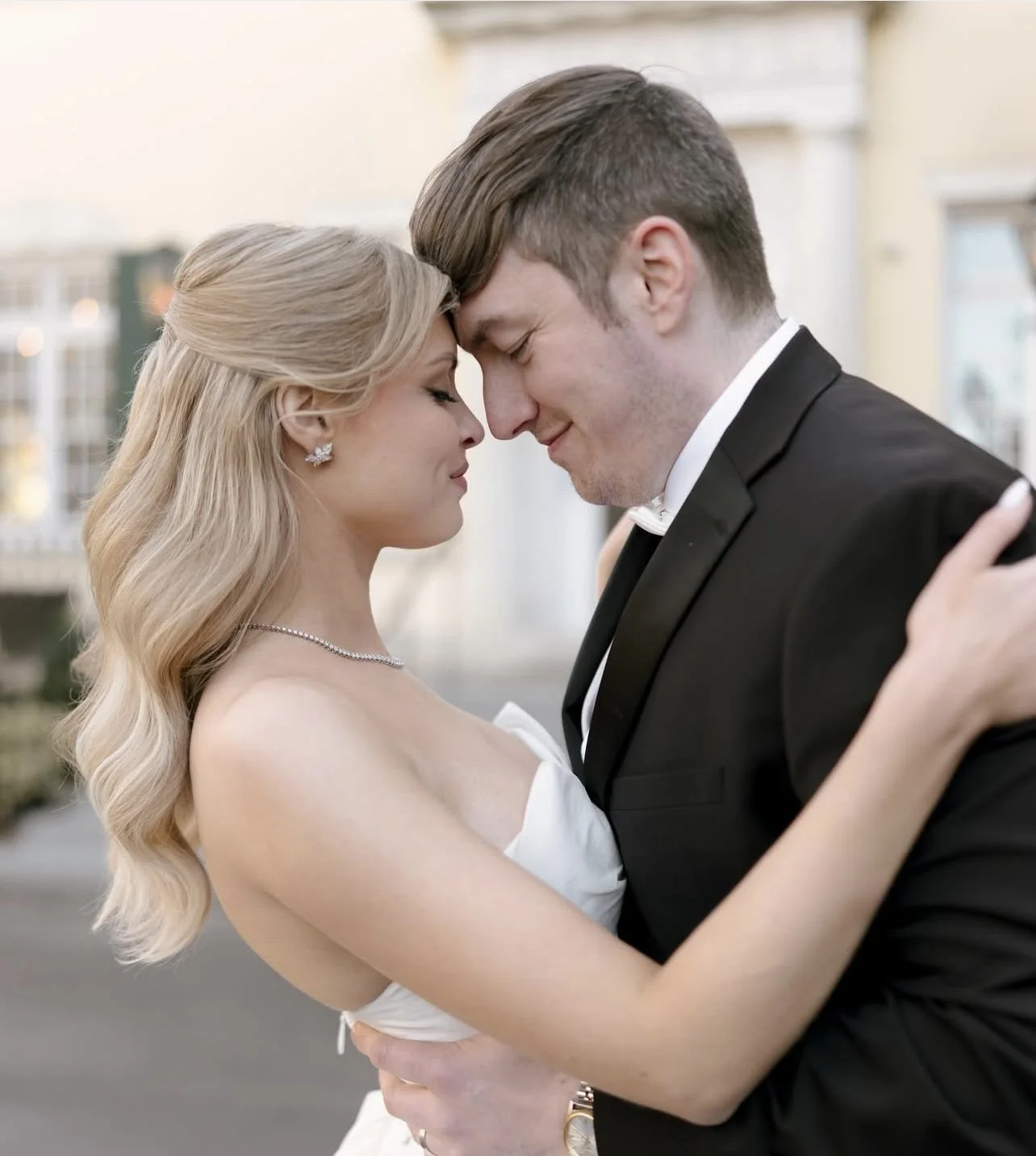 A bride and groom embrace in wedding attire with their foreheads touching, eyes closed, smiling, outdoors with blurred buildings in the background.