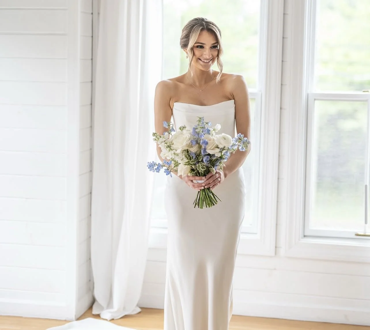 A smiling bride in a white strapless wedding dress holding a bouquet of white and light purple flowers, standing in a bright room with white walls and windows.