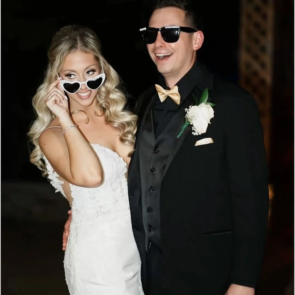 A bride and groom at a wedding, both wearing sunglasses and smiling, with the bride holding heart-shaped glasses near her face. The groom is in a black tuxedo with a gold bow tie and a white flower boutonniere.