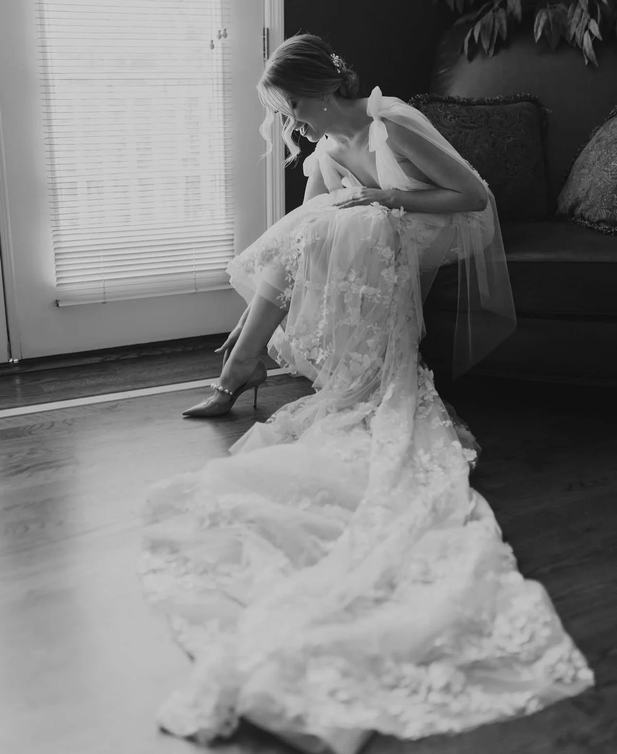 A bride in a wedding dress putting on high heel shoes, sitting on a sofa near a door with blinds, in a black and white photo.
