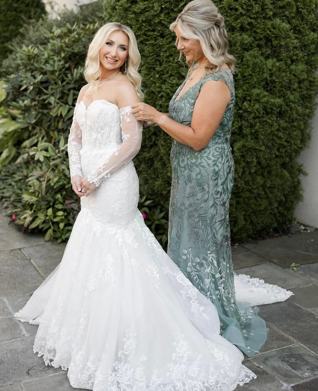 A bride in a white lace wedding dress smiling as an older woman helps her with her dress. They are outdoors with green bushes in the background.