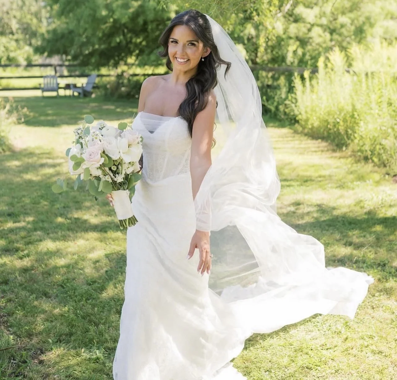 A smiling bride with long dark hair, wearing a white wedding dress and veil, holding a bouquet of white and pink flowers, standing outdoors on a sunny day with greenery in the background.