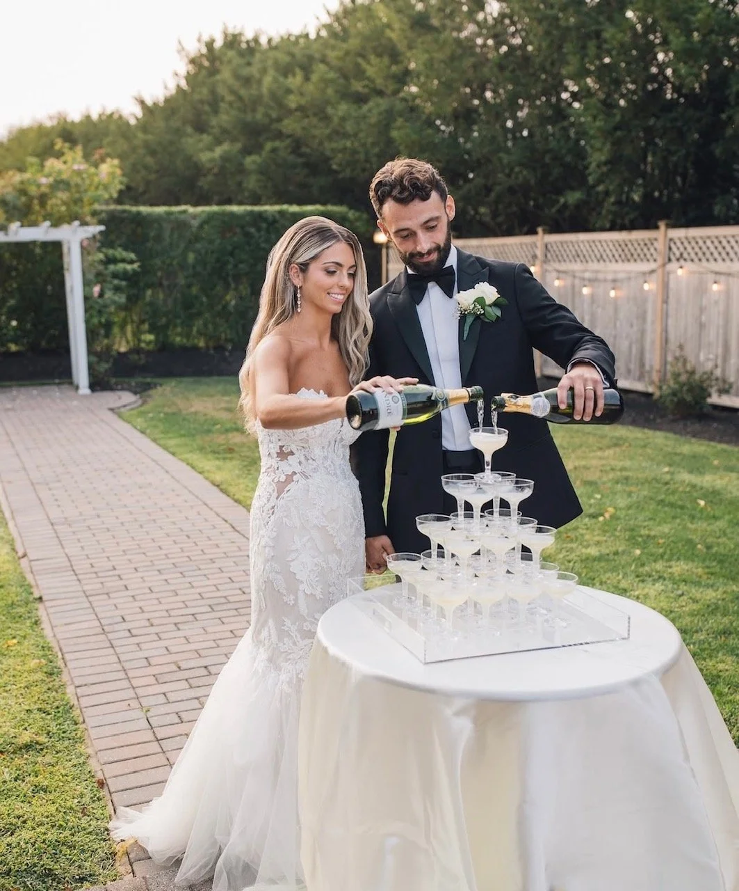 Bride and groom pouring champagne into a pyramid of glasses during their wedding celebration outdoors.