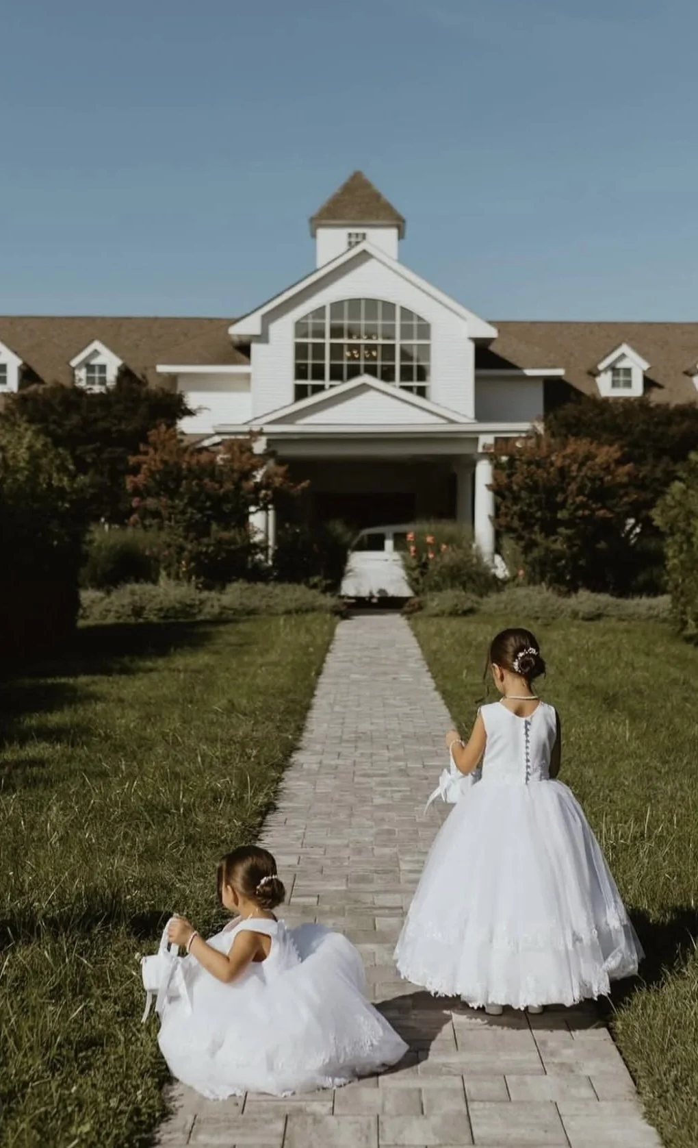 Two young girls dressed in white dresses walk on a stone path toward a large white house with a peaked roof and large window, surrounded by greenery and trees.